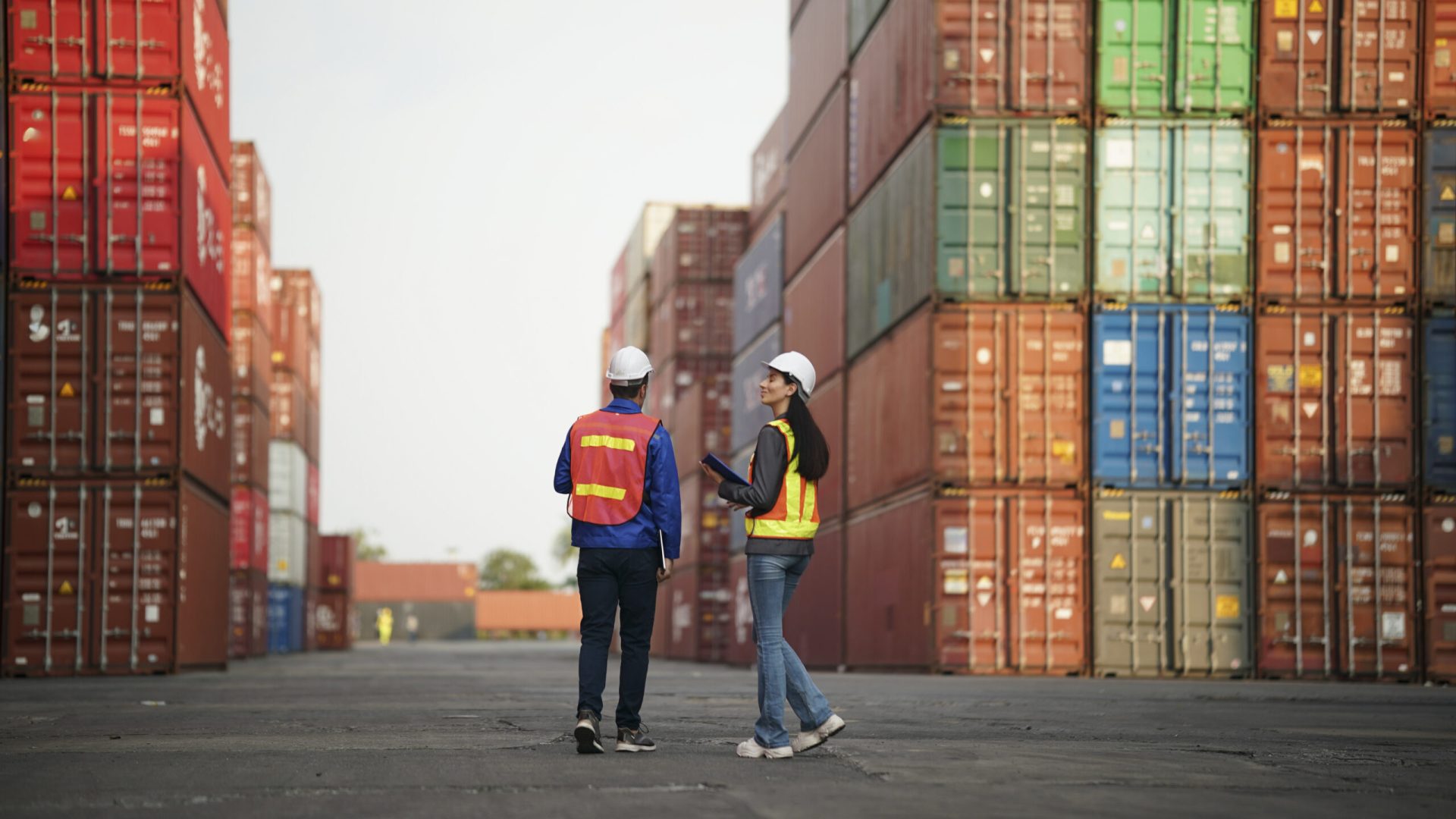 Two workers in safety gear review plans and discuss operations in a container yard.